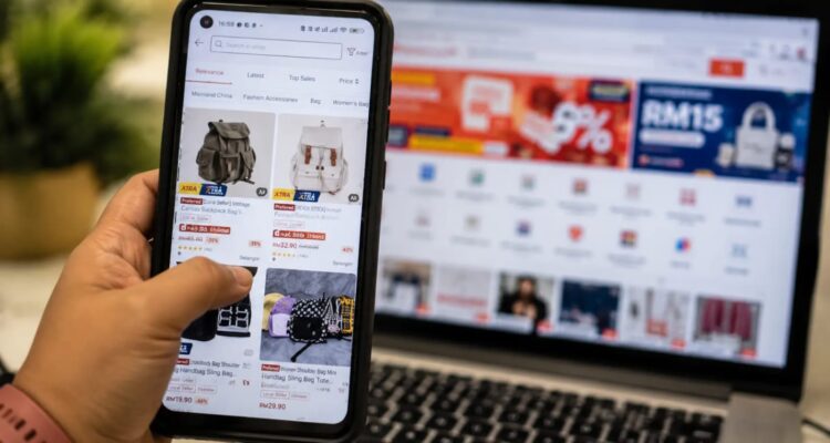 Pelaku UMKM menggunakan smartphone untuk mengelola penjualan di marketplace dengan laptop di latar belakang.
